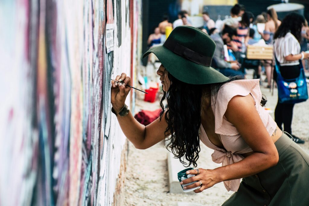 Young woman wearing hat painting a mural on an urban street wall during the day.